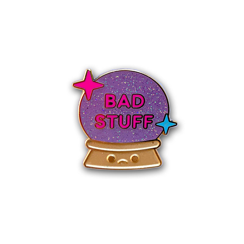 Gold pin of a crystal ball with a sad face on the base and purple sparkles with Bad Stuff written in pink on the ball.