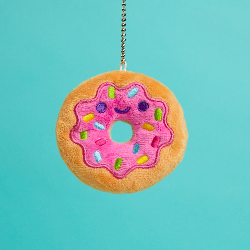 A small plush donut with pink frosting and sprinkles on it that is smiling on a chain. 